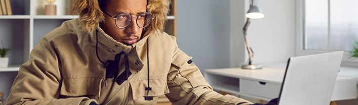 Freezing man in warm jacket and gloves working at computer in office