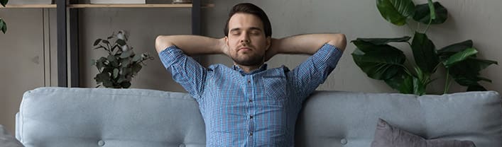 Calm man sit on sofa with eyes closed put hands behind head stretching on soft cozy sofa in living room