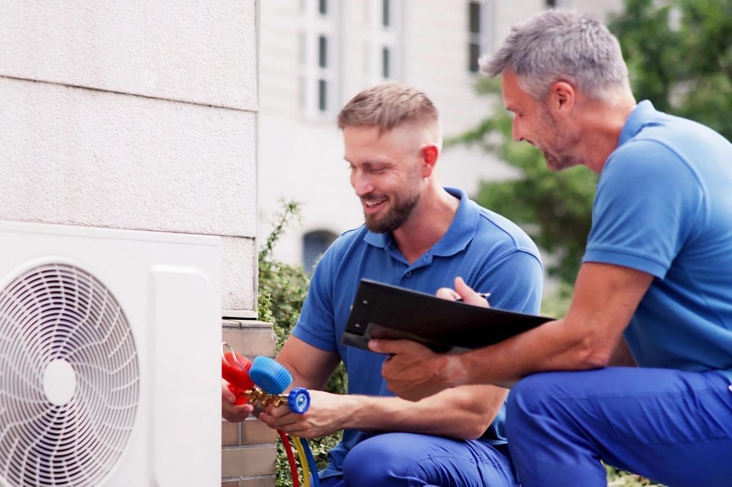 two HVAC technicians working on an outdoor heat pump unit.